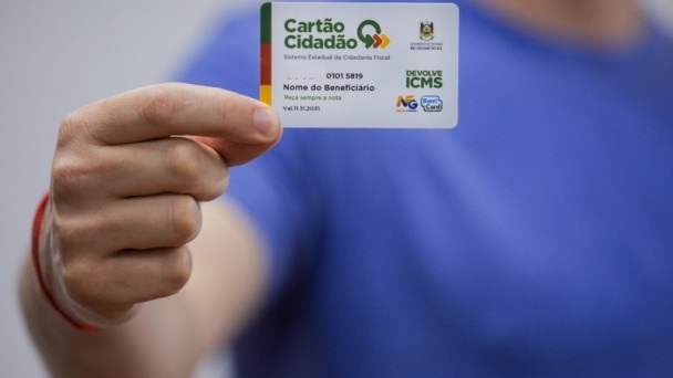 Mão segura Cartão Cidadão. Devolve ICMS enquanto pessoa de camiseta azul posa em fundo neutro