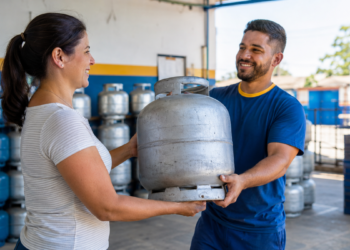 Mulher recebe botijão de gás de funcionário sorridente em distribuidora com vários cilindros ao fundo