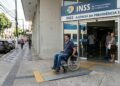 Homem em cadeira de rodas utiliza rampa acessível na entrada da agência INSS com pessoas ao redor