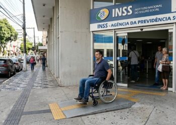 Homem em cadeira de rodas utiliza rampa acessível na entrada da agência INSS com pessoas ao redor