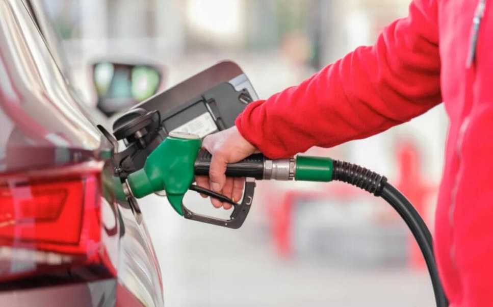 Mão abastecendo carro vermelho com bomba de combustível verde em posto, pessoa usa jaqueta vermelha
