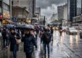 Pedestres com guarda-chuvas andam em rua movimentada de São Paulo sob chuva intensa e céu nublado