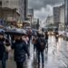 Pedestres com guarda-chuvas andam em rua movimentada de São Paulo sob chuva intensa e céu nublado