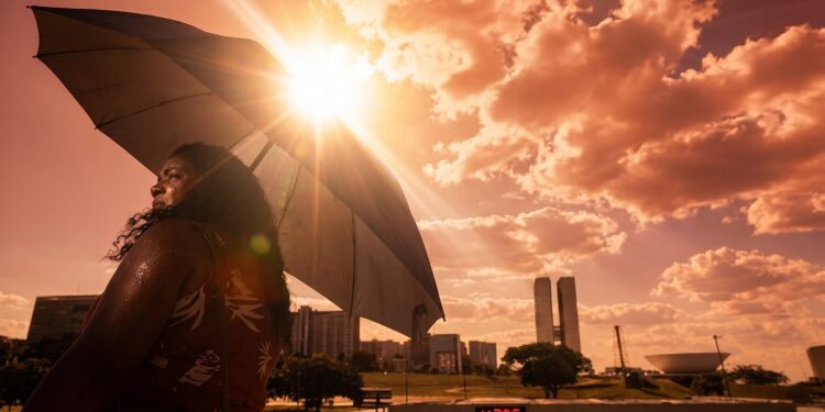 Mulher negra segura guarda-chuva sob sol intenso com termômetro marcando 43°C e Congresso Nacional ao fundo em Brasília