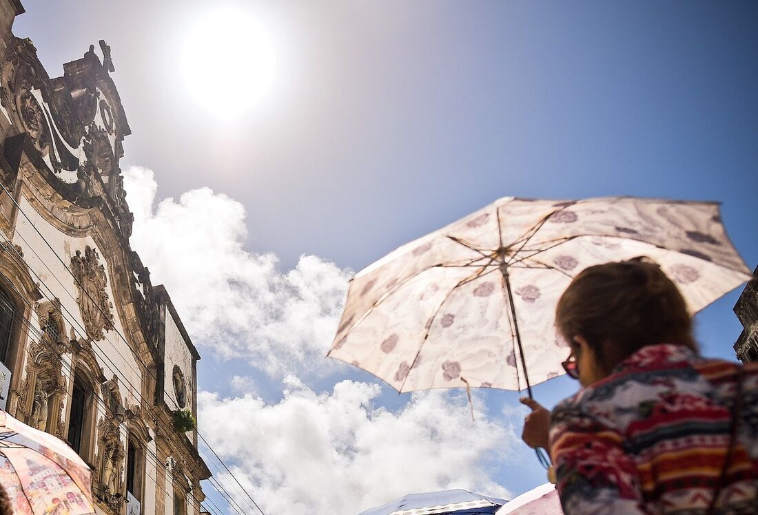Pessoa segura sombrinha estampada sob céu azul e sol forte, com fachada barroca de igreja colonial ao fundo