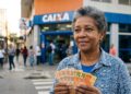 Mulher idosa segura notas de real em frente à agência da Caixa, sorrindo levemente