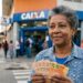 Mulher idosa segura notas de real em frente à agência da Caixa, sorrindo levemente