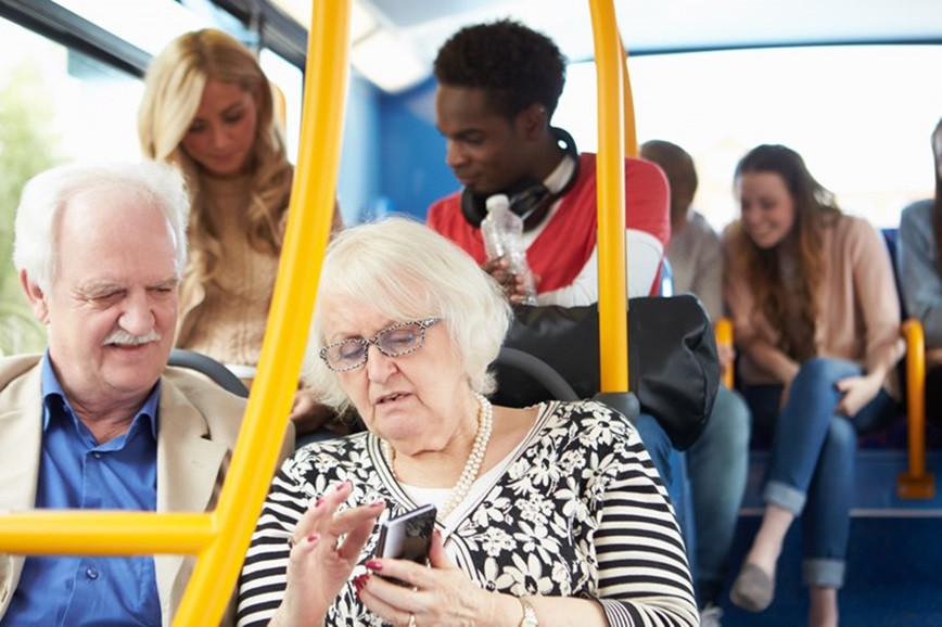 Casal idoso sentado em ônibus observa celular enquanto outros passageiros conversam ao fundo