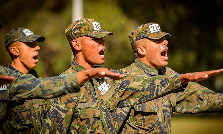 Três jovens soldados com farda camuflada e boné fazem juramento com o braço estendido durante o treinamento.