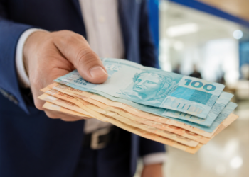 Mão masculina entrega várias cédulas de reais em frente a fundo desfocado, foco em notas azuis e laranjas