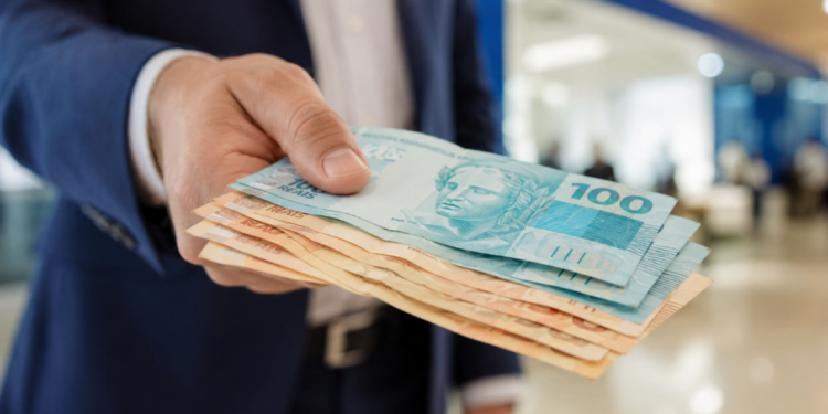 Mão masculina entrega várias cédulas de reais em frente a fundo desfocado, foco em notas azuis e laranjas