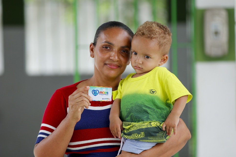 Mulher com camisa listrada segura criança e exibe cartão do programa Mães de Pernambuco em cenário institucional