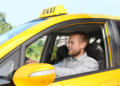 Motorista de táxi sorridente conduzindo veículo amarelo em área urbana.