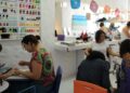 Cinco mulheres em salão de beleza fazendo unhas e cabelos, esmaltes coloridos na parede ao fundo