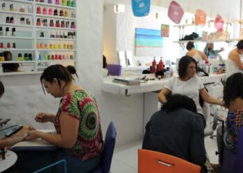 Cinco mulheres em salão de beleza fazendo unhas e cabelos, esmaltes coloridos na parede ao fundo