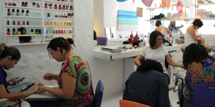 Cinco mulheres em salão de beleza fazendo unhas e cabelos, esmaltes coloridos na parede ao fundo