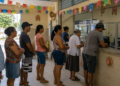 Sete pessoas de diferentes idades aguardam em fila em agência com decoração de festa junina
