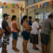 Sete pessoas de diferentes idades aguardam em fila em agência com decoração de festa junina