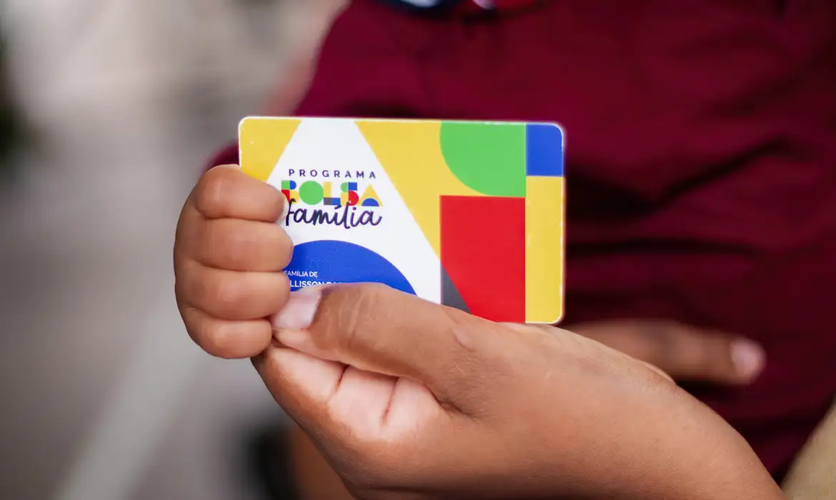 Mãos de adulto e criança segurando cartão colorido do Programa Bolsa Família