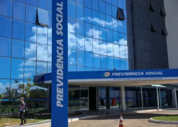 Fachada espelhada do prédio do INSS Previdência Social com céu azul refletido e mulher caminhando