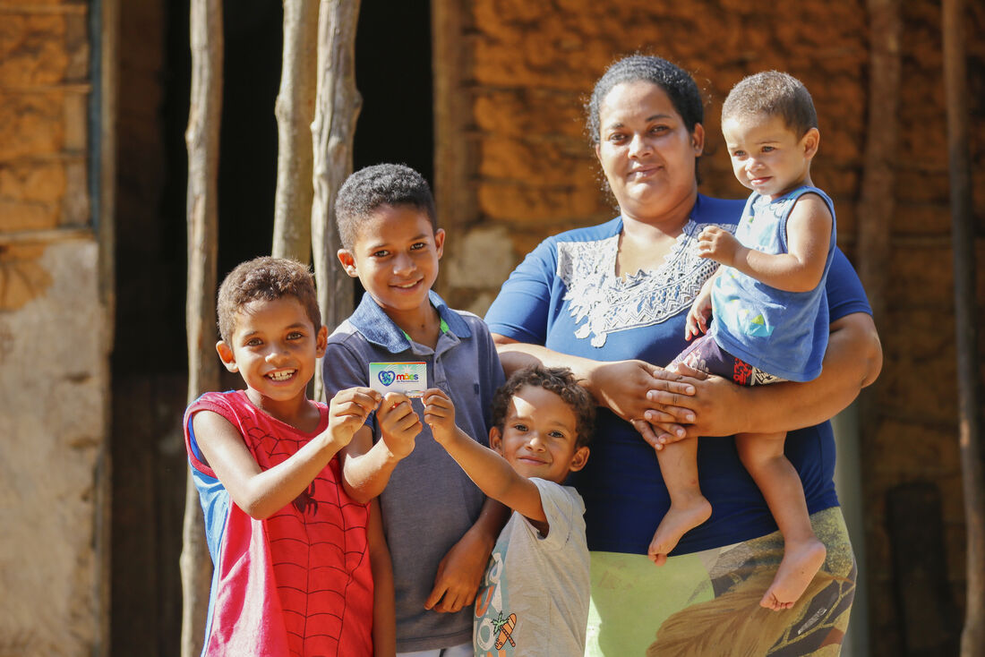 Mulher com quatro crianças sorri enquanto exibe cartão Mães de Pernambuco na frente de casa simples