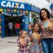 Mulher e duas meninas sorrindo, segurando notas de real, em frente à Caixa decorada para São João