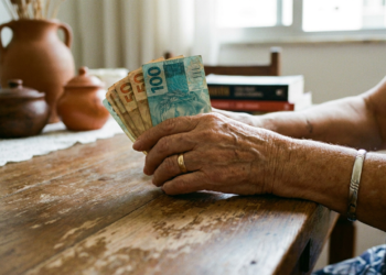 Mãos de idosa seguram notas de 100 e 50 reais em ambiente doméstico.