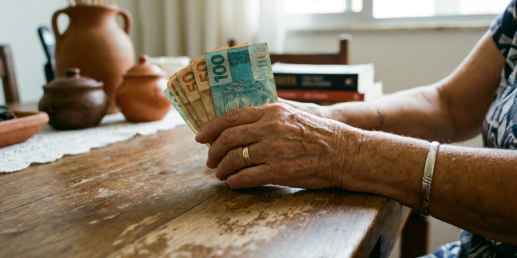 Mãos de idosa seguram notas de 100 e 50 reais em ambiente doméstico.