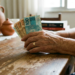 Mãos de idosa seguram notas de 100 e 50 reais em ambiente doméstico.
