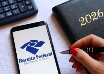 Celular com logo da Receita Federal, calculadora e agenda preta de 2026 ao lado de mão segurando caneta.