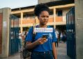 Jovem negra de uniforme azul lê mensagem no celular informando bloqueio temporário do benefício escolar