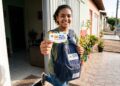 Jovem mostra cartão do Pé-de-Meia sorrindo, com mochila azul do programa, em frente à casa