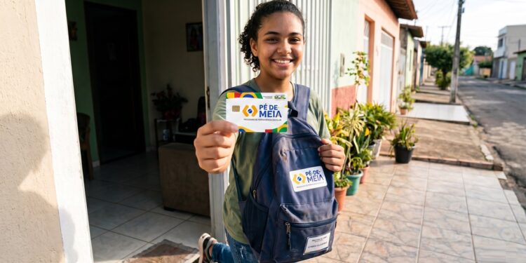 Jovem mostra cartão do Pé-de-Meia sorrindo, com mochila azul do programa, em frente à casa
