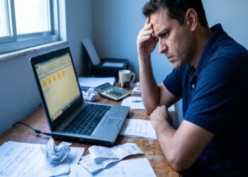 Homem de camisa polo azul-marinho apoia a cabeça na mão enquanto olha para o laptop cercado por papéis e calculadora