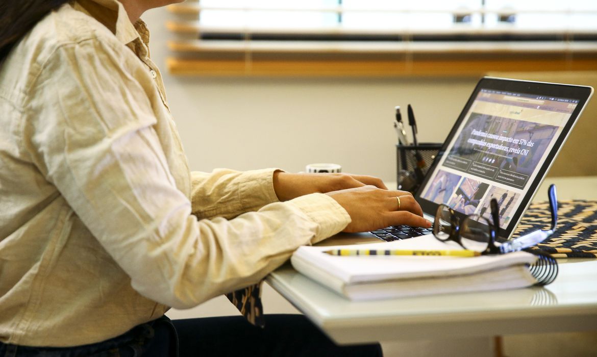 Mulher de camisa bege digitando em notebook, óculos e caderno na mesa ao lado