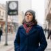 Mulher com gorro cinza e cachecol vermelho caminha em calçadão urbano durante dia frio, relógio marca 14°C