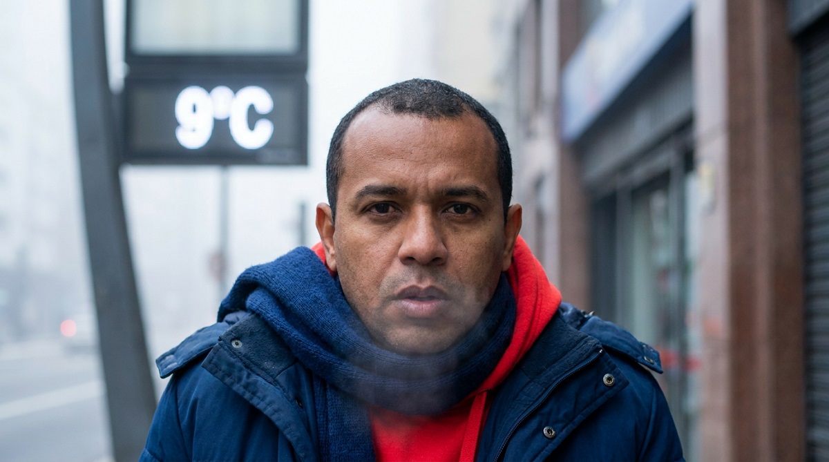 Homem negro de jaqueta azul e moletom vermelho em rua com display eletrônico mostrando 9°C ao fundo