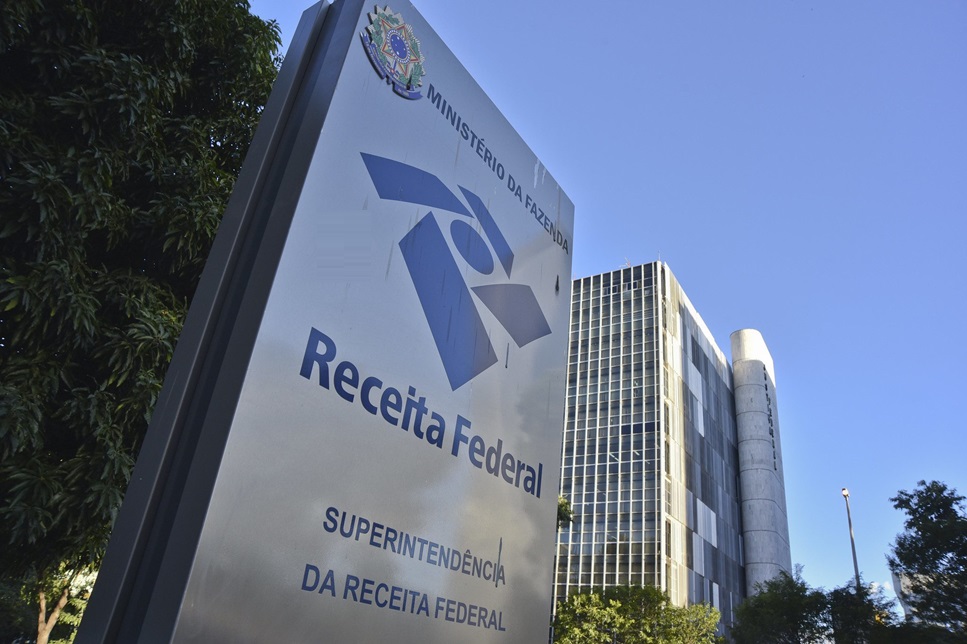 Placa da Receita Federal com edifício moderno ao fundo e céu azul, destacando texto e logotipo oficiais