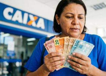 Mulher segura notas de 10, 20, 50 e 100 reais em frente à agência da Caixa Econômica Federal