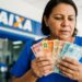 Mulher segura notas de 10, 20, 50 e 100 reais em frente à agência da Caixa Econômica Federal