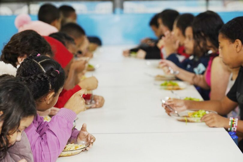 Crianças sentadas lado a lado em mesa de refeitório escolar, comendo almoço