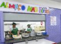 Duas merendeiras servindo comida ao balcão sob faixa “Almoço nas Férias” em refeitório escolar