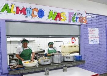 Duas merendeiras servindo comida ao balcão sob faixa “Almoço nas Férias” em refeitório escolar