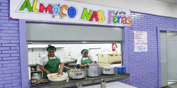 Duas merendeiras servindo comida ao balcão sob faixa “Almoço nas Férias” em refeitório escolar