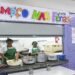Duas merendeiras servindo comida ao balcão sob faixa “Almoço nas Férias” em refeitório escolar