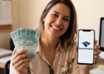 Mulher sorrindo segura notas de R$100 e celular com logo da Receita Federal em escritório.