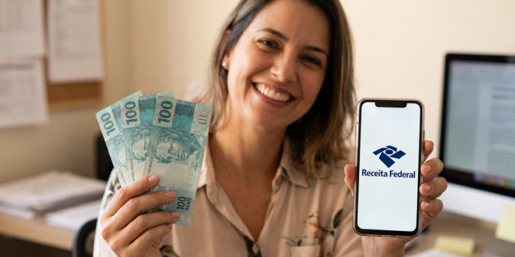 Mulher sorrindo segura notas de R$100 e celular com logo da Receita Federal em escritório.