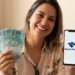 Mulher sorrindo segura notas de R$100 e celular com logo da Receita Federal em escritório.