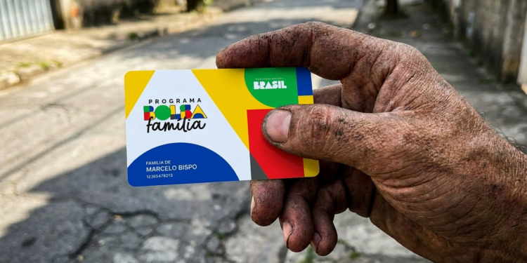 Mão suja exibe cartão do Bolsa Família em rua simples do Brasil.