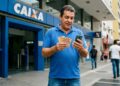 Trabalhador confere o celular e segura cédulas em frente à Caixa, em referência ao Abono Salarial de junho de 2026